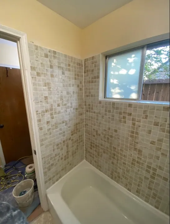 White tub beneath a wide sliding window, surrounded by neutral small square wall tile with pale yellow-beige walls above.