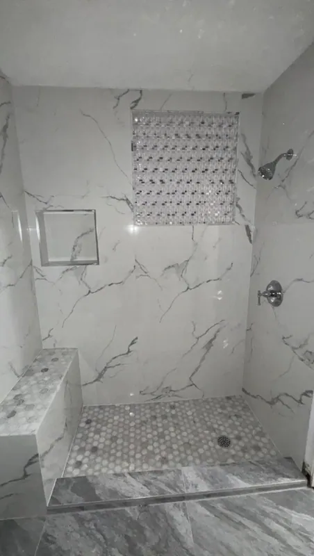 Shower with white gray-veined wall panels, a built-in bench, a mosaic feature inset, and a rounded pebble floor.